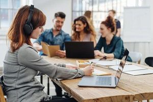 A woman wearing headphones works on a laptop at a shared table while three colleagues collaborate nearby with laptops and documents.
