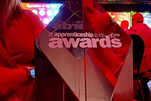 Photograph of a reflective, pyramid-shaped trophy with "apprenticeship guide awards 2026" etched on its surface, held by a person in a red outfit. Background shows a dimly lit event space with colorful lights and seated attendees, indicating an awards ceremony setting.