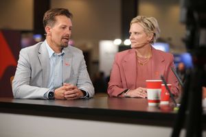 Two speakers wearing microphones participate in a live interview at a broadcast desk, discussing enterprise technology topics in a studio setting with a blurred event backdrop.