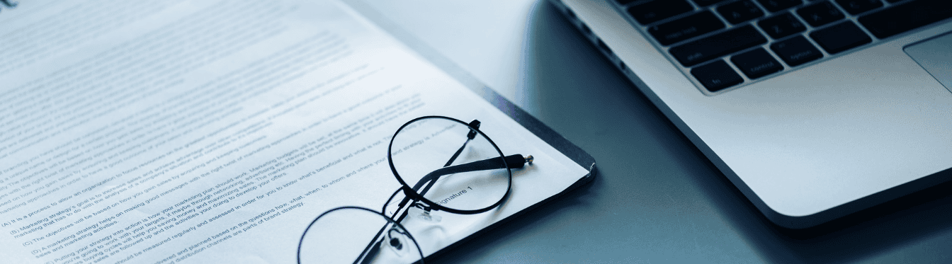A pair of glasses resting on a printed contract sitting on a desk next to an open laptop