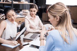 Three professional women sitting around a table, with one writing on a notepad.