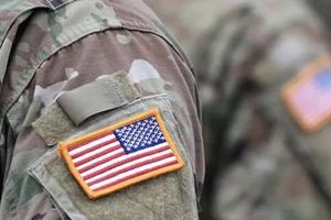 Close-up of a U.S. flag patch on the sleeve of a camouflage military uniform, with another uniformed service member blurred in the background.