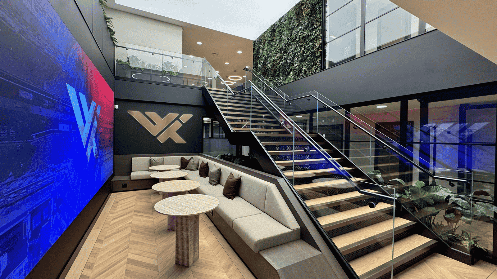 Photograph of a modern office lounge area featuring a large digital screen with blue and red graphics on one wall and a wooden logo on another. The space includes a long beige sofa with brown cushions, round tables, a glass staircase with black railings, and a vertical green plant wall.