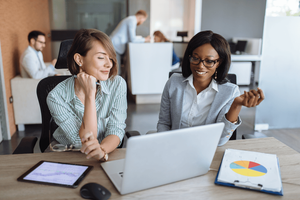 Two professional business women sitting at a desk, smiling at an open laptop as if they just read WWT's article on the four steps for turning an AI use case into a running solution and are pleased.