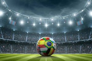 Soccer ball featuring multiple national flags positioned on a stadium field under bright lights, with a packed crowd in the background, representing international competition and global sports.