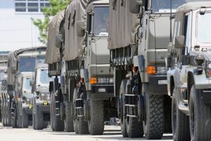 A line of military transport trucks with canvas-covered cargo areas is parked along a roadway, stretching into the distance in an outdoor industrial setting.