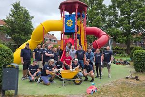 Team of volunteers wearing matching shirts pose in front of a colorful playground structure with red and yellow slides, gathered around a wheelbarrow during a community service project in a neighborhood park.