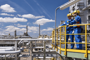 Two two workers in blue jumpsuits and yellow hardhats standing on a platform overlooking an oil and gas operation. 
