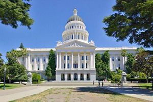 California State Capitol building with its white domed facade centered between large trees, surrounded by landscaped grounds and walkways under a clear blue sky.