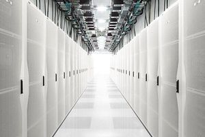 Long corridor of a modern data center lined with white server racks, with overhead cabling and bright lighting creating a symmetrical view toward the far end of the room.
