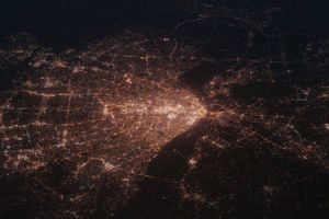 City streetlights and illuminated roadways form a dense web of glowing connections across a metropolitan area at night, viewed from above against a dark background.