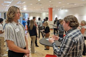 A volunteer talks with a high school student wearing a name tag at a networking event, with other attendees mingling in the background.