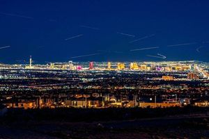 Las Vegas skyline illuminated at night, viewed from a nearby hillside neighborhood with aircraft light trails streaking across the sky.