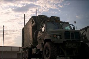 Military-style tactical truck with a large enclosed equipment module parked outdoors, photographed from a low angle against a partly cloudy sky, emphasizing its rugged tires, reinforced chassis, and mobile communications or command capability.