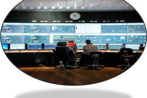 Two operators seated at desks facing a wall of large monitors in an industrial control room