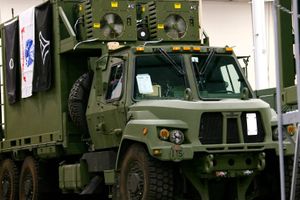 Military-style tactical vehicle equipped with large communications or power units displayed indoors, with service flags mounted along the side and heavy-duty off-road tires visible.  Provide your feedback on BizChat