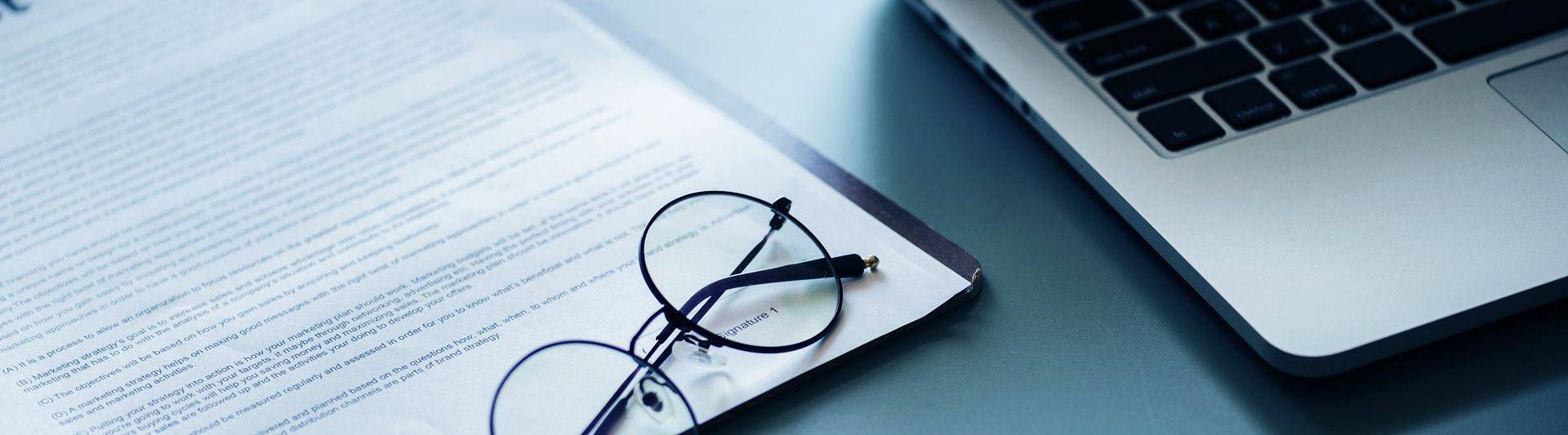 Pair of glasses resting on a printed contract next to a laptop. 