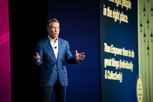 Jim Kavanaugh in a blue checkered blazer delivers a presentation on a conference stage beside a large screen displaying a message about empowering others to achieve great things professionally and collectively.