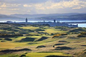 Rolling green fairways and rugged dunes stretch toward a distant coastal town and shoreline, with rolling hills and soft sunlight beneath a cloud-filled sky.