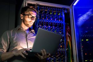 A person uses a laptop beside illuminated server racks filled with network cables and equipment.