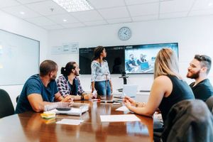 A team gathers around a conference room table while one person presents beside video conferencing screens displaying remote participants.
