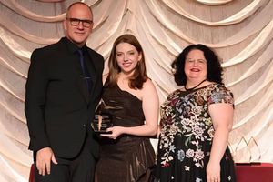 Three people pose together at an awards event, with the person in the center holding a trophy in front of a draped backdrop.